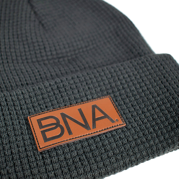 Close up of Charcoal gray waffle knit beanie with brown faux leather patch on cuff.  Patch features debossed BNA logo.