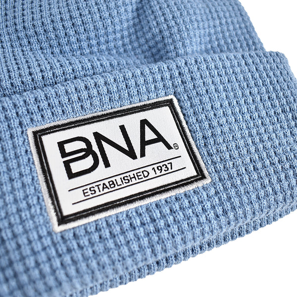 Close up of Light blue waffle knit beanie with white woven label on cuff.  Label features black border and BNA logo above "established 1937".
