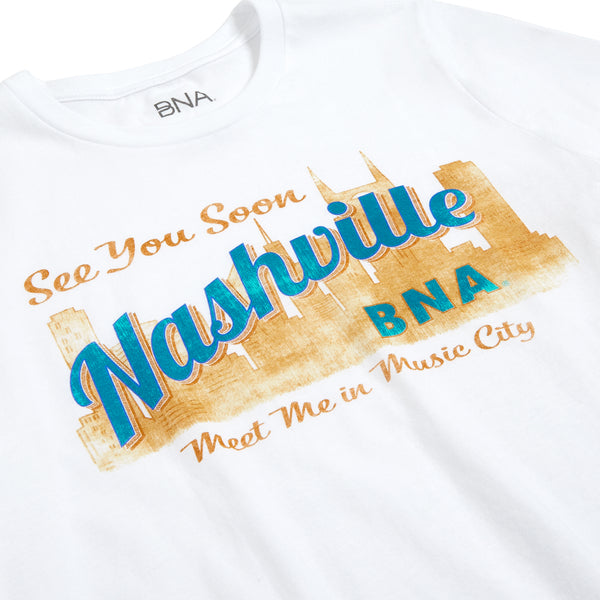 Detail shot of the Vintage Postcard Tee print.  Light tan and teal design looks like a vintage postcard with "See You Soon" in a script font printed above an illustration of the Nashville Skyline. "Nashville" is superimposed over the skyline, with the tag line "Meet me in Music City" below.