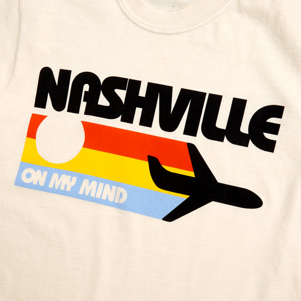 detailed view of front graphic depicting a black plane and three vintage stripes in orange, yellow, and light blue.  Tee reads "Nashville on my mind" in retro bubble letters.  T-shirt is cream.