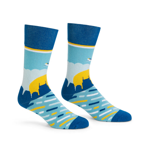 Light blue socks with a navy cuff, toe and heel show the Nashville skyline at sunrise.  Dark blue, white, and yellow striping across the top of the foot and the BNA logo on the bottom of the foot. An aircraft is seen mid air above clouds on the front of the socks.