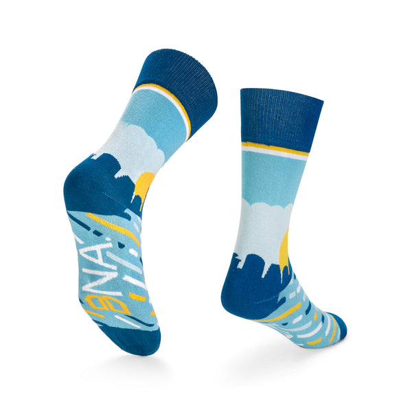 Back view of light blue socks with a navy cuff, toe and heel show the Nashville skyline at sunrise.  Dark blue, white, and yellow striping across the top of the foot and the BNA logo on the bottom of the foot. 