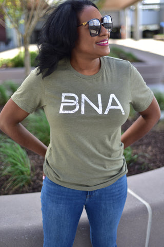 A female model wears the BNA Logo T-Shirt in Heather Olive outdoors at BNA.