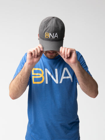 Man wearing BNA logo cap in gray.  Cap has the BNA logo in a 3D embroidery (gold infinity B with white NA). 