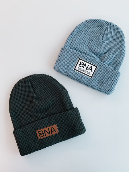 Image shows a light blue waffle knit beanie that is cuffed with a white woven label. Label says " BNA" and "established 1937" in black writing. Below it is a cuffed charcoal waffle knit beanie wit ha brown faux leather patch depicting the BNA logo.